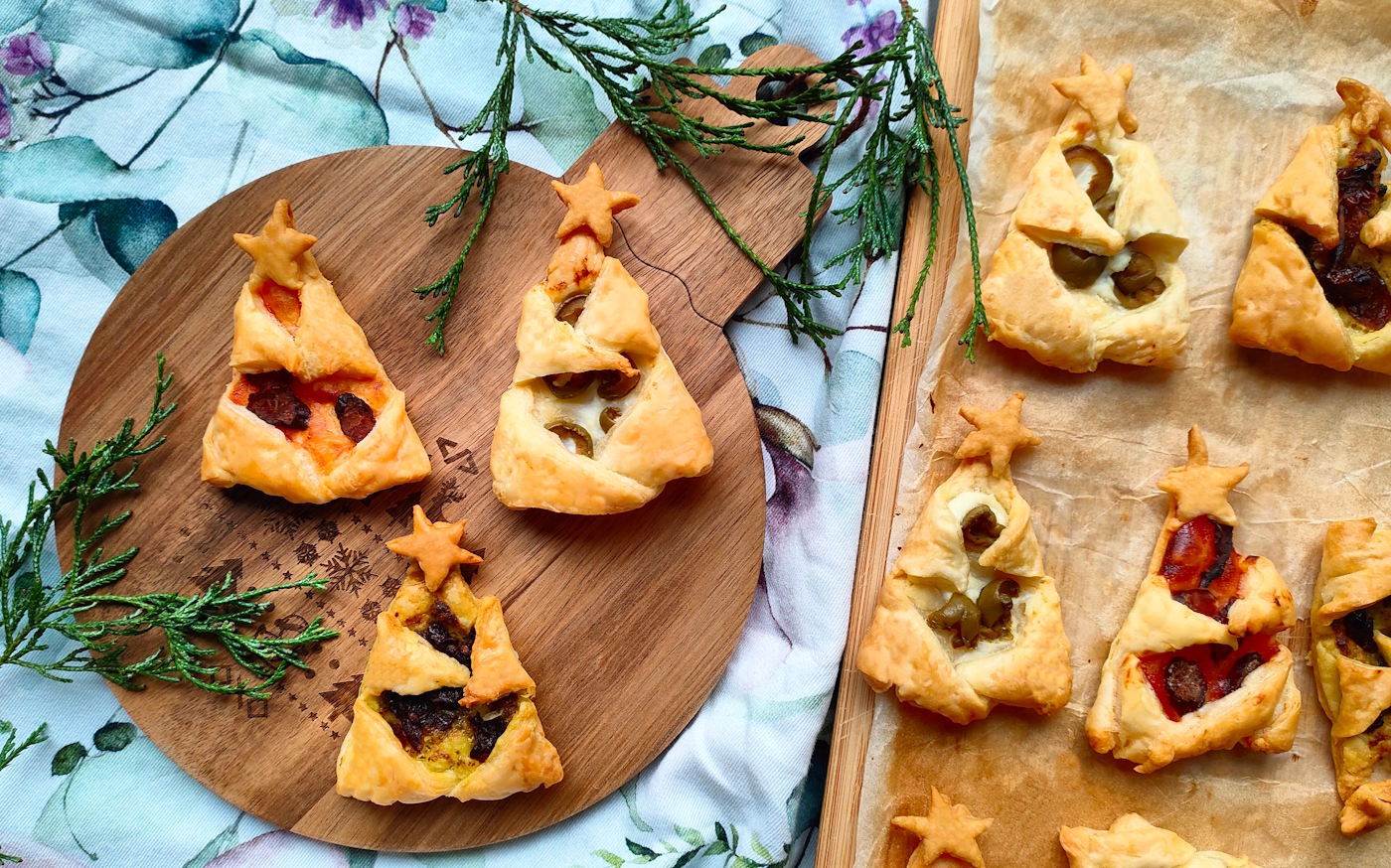 Wytrawne choinki z ciasta francuskiego z pesto, oliwkami, kabanosem i pomidorami suszonymi, ułożone na desce i papierze do pieczenia.